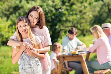 Obraz premium Mother hugging daughter at a sunny family picnic with relatives enjoying an outdoor meal and relaxed multigenerational gathering