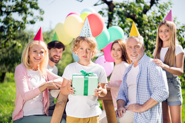 Fototapeta premium Multi-generation family celebrating a child's birthday outdoors with balloons, party hats and the child holding a wrapped gift at a sunny backyard party
