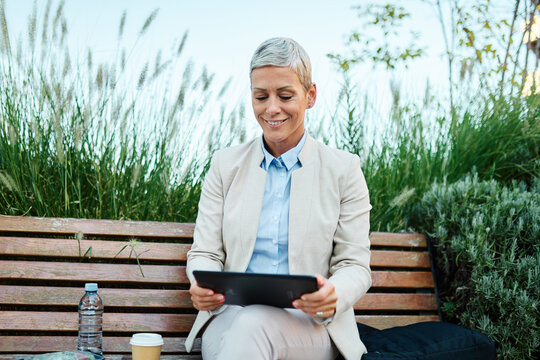 Portrait of a young businesswoman woman using a tablet or laptop computer and sitting on a bench in a city park, having a break for a coffee and water drink, surrounded by modern corporate office buil