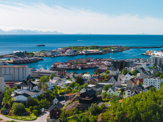 Naklejka premium Aerial view of a Lofoten Islands coastal town in Norway with wooden houses, turquoise harbor, small fishing boats, a rocky islet marina, and distant mountain silhouettes.