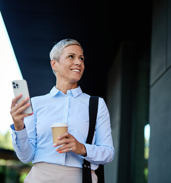 Portrait of a young businesswoman woman using a smartphone mobile phone and holding a cup of coffee walking down the street, surrounded by moder corporate office buildings