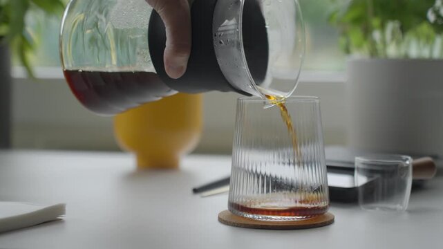 Brewed coffee is poured from glass carafe into drinking glass on cork coaster. Concept of coffee serving, fresh brew and morning drink