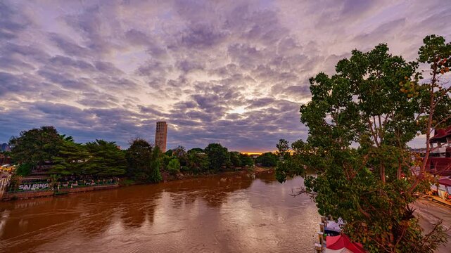 Time lapse A dramatic cloudy sky casts soft light over a calm river at sunset, with reflections shimmering on the water and lush greenery lining the riverbank at Ping river in Chiang Mai