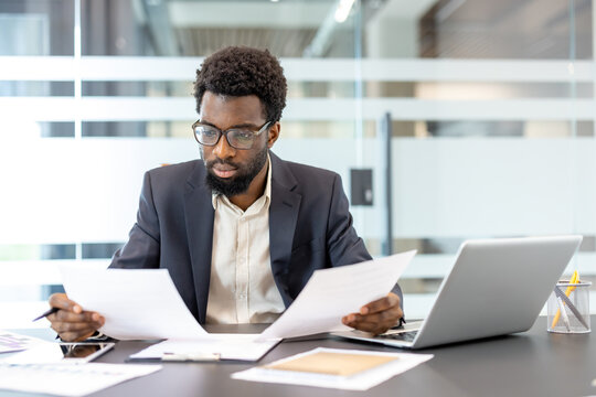 Young businessman wearing a suit and glasses sitting at a modern office desk, meticulously reviewing important paperwork and reports while using a laptop to analyze data