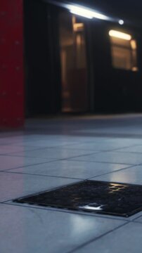 A person walks in a hallway with glossy tiles. A drain cover is visible on the floor. The walls are dark with a red column. Natural light comes in from a nearby window.