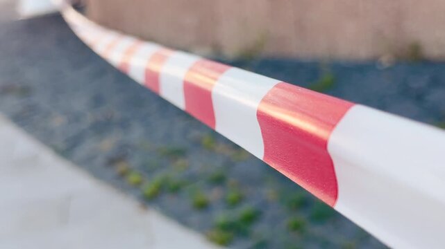Red and white caution tape stretches tightly across a paved outdoor walkway. This tense barrier blocks the path, bringing an uneasy feeling of anxiety and curious worry.