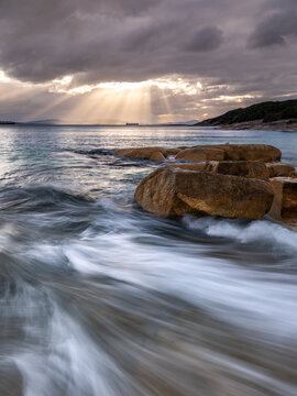 View of Whalers Cove with large granite boulders in the foreground and ocean waves washing over the shore under a dramatic cloudy sky with sun rays in Albany, WA, Australia.