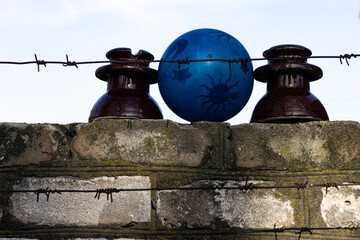 Fototapeta premium Blue ball with sun pattern between ceramic electrical insulators on a brick wall behind barbed wire. The shot has conceptual and symbolic meaning, barbed wire and bright ball with sun and star pattern
