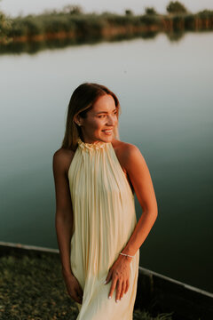 Woman stands by a lake during sunset wearing a light dress and smiling