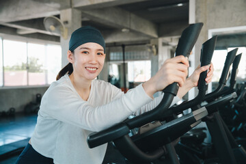 Fototapeta premium Woman gym exercise fitness elliptical machine smile indoor workout cardio, happy woman exercising elliptical machine indoor gym, wearing headband and white shirt, holding handle during cardio
