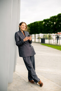 Woman in business suit using phone while leaning against a wall outdoors near green trees