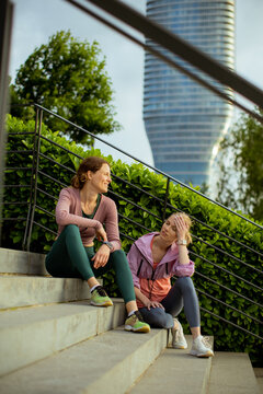 Two women sit on steps and talk near a tall building in the city during the day