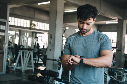 Man gym watch fitness exercise athlete checking wearable device in industrial training center with dumbbell equipment focused expression