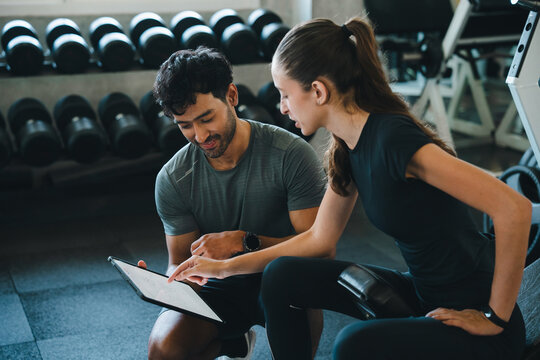 Personal trainer gym woman man clipboard workout consultation fitness coaching exercise, focused adult pair reviewing training plan near dumbbell rack on bench