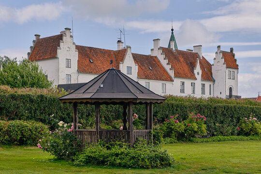 View of the historic Bosjokloster Abbey with white walls and red-tiled roof, featuring a wooden gazebo in the foreground and a lush green garden in Bosjokloster, Skane County, Sweden.