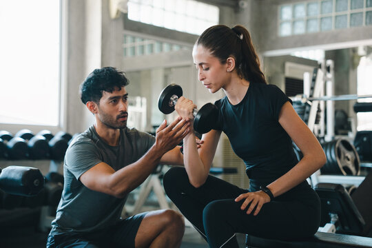 Fitness woman trainer dumbbell gym exercise coaching strength workout support, woman lifting dumbbell with male trainer guidance modern gym, focused expression during strength exercise