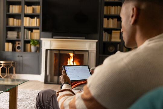 African American man sitting on sofa holding tablet, facing lit fireplace with TV, copy space