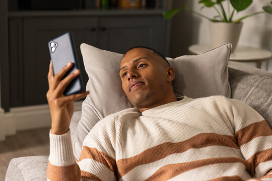 Man reclining on light-gray sofa holding smartphone near face and wearing striped sweater