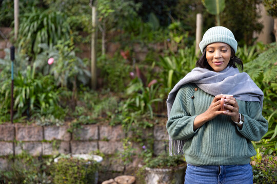 Woman standing right side garden holding ceramic mug with beanie and scarf, copy space