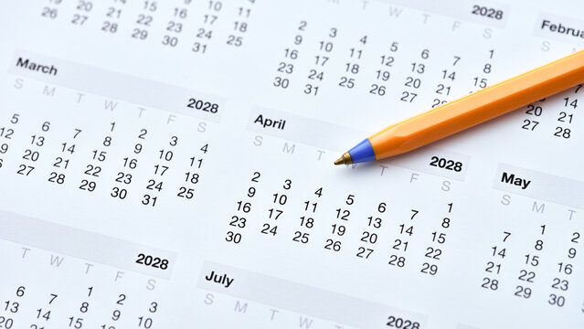 An orange ballpoint pen resting on a printed paper calendar for the year 2028. Close up.