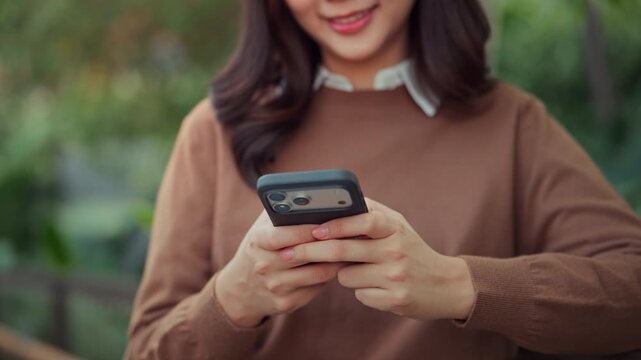 Close up of happy Asian woman typing text message on smartphone outdoors, smiling female using mobile phone for chatting and online shopping in city park, connection, technology, and modern lifestyle