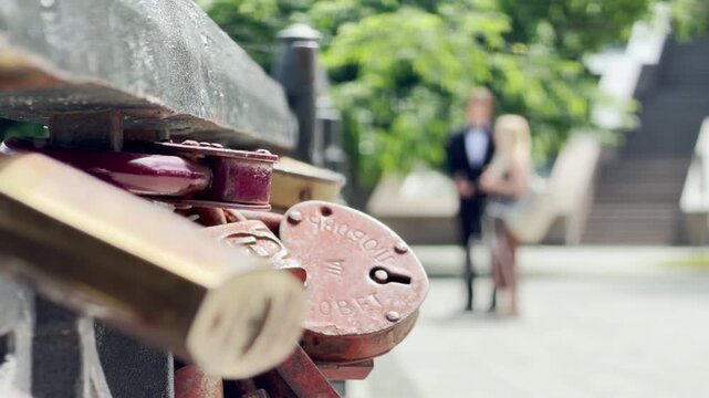 A rusty heart padlock hangs on a metal bridge railing. In the blurry background, a happy newlywed couple stands together in a sunny park. This lovely view brings a deeply romantic feeling.