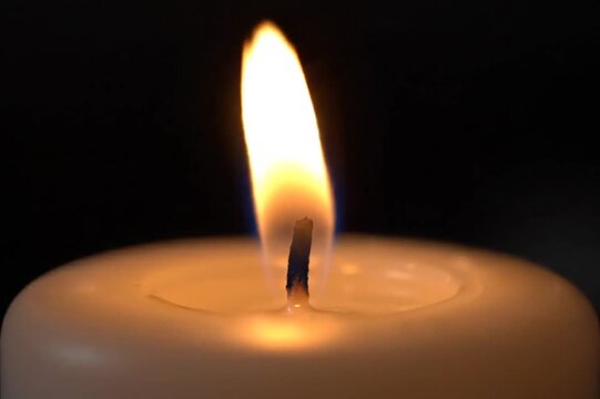 Close-up macro shot of a white candle wick burning with a bright yellow and orange flame against a dark black background, showcasing the molten wax