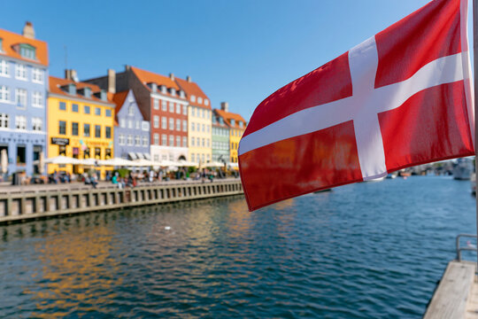 Red Danish flag waves gently in the breeze. Colorful buildings line a calm waterfront canal. Sunlight glints off the rippling water surface. Quiet, picturesque scene invites travelers