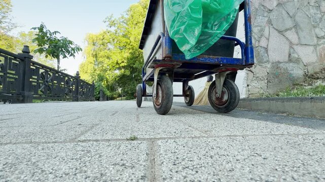 A blue utility cart sits quietly on a sunny paved path in Simferopol, Crimea. A straw broom rests nearby in the peaceful park. The empty walkway feels serene and orderly.