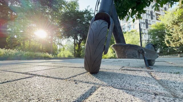 An electric scooter stands on a paved path in Simferopol, Crimea. Bright morning sunbeams flash through the park trees, creating a warm, joyful, and serene atmosphere.