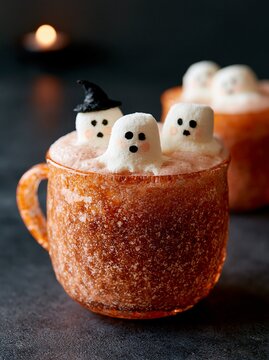 Textured orange speckled glass mug filled with a festive drink topped with marshmallow ghosts wearing hats, set against a dark background with a candle