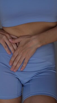 Young woman holds her side and lower abdomen, showing pain after exercise, muscle strain, cramps, digestive discomfort or possible women&rsquo;s health issue in a body wellness concept on a grey background 