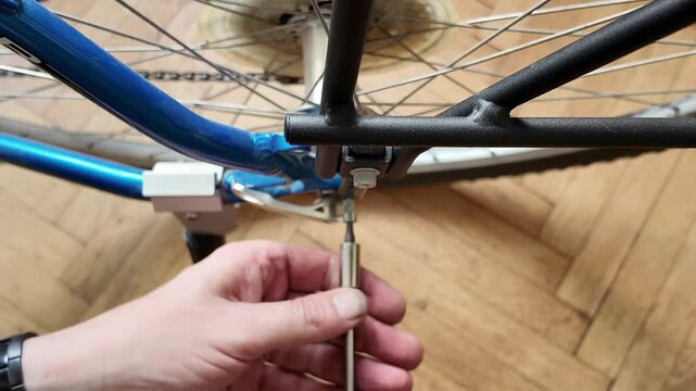 Close-up of hands disassembling a bicycle frame with tools on a wooden floor. A person's hands use a screwdriver to work on the blue frame of a bicycle, with spokes and chain visible against a wood