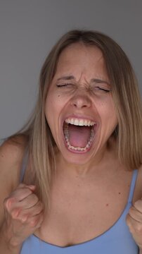Young blonde woman screams loudly with intense expression, releasing built-up emotions, stress, anger and frustration, showing emotional outburst, catharsis and freedom on a grey background