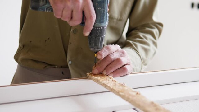 Close view shows worker hands finishing drilling fastener into rough wooden plank on white workbench, with centered action, bright indoor background, and visible wood dust