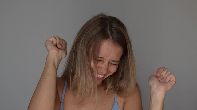 Young blonde woman claps her hands and gestures with excitement, expressing joyful anticipation, happiness and playful enthusiasm in a lively emotional reaction concept on a grey background