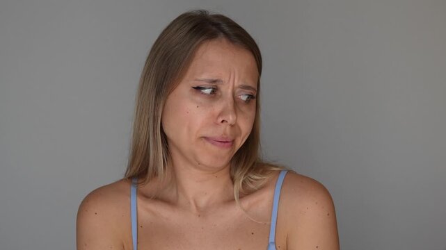 Young blonde woman smells her underarm and reacts with unpleasant emotion, showing bad armpit odor, body smell and hygiene concern in a funny lifestyle concept on a grey background