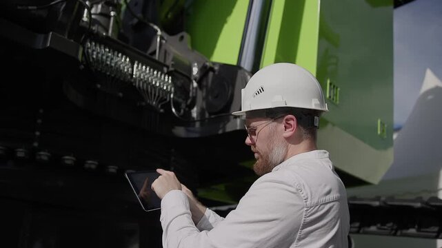 Worker mechanic checks hydraulic hoses of excavator with tablet computer. Man in hard hat industrial worker.
