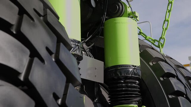 Truck wheel and hydraulic suspension close up in outdoor farm machinery yard showing rotating tire tread and green cylinder detail.