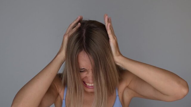 Young blonde woman hits or grabs her head in frustration, expressing self-blame, regret and the feeling of &ldquo;what have I done&rdquo; in an emotional lifestyle concept on a grey background 