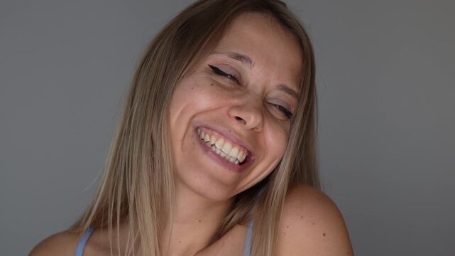 Young blonde woman gives a flirty sideways look with a mysterious smile, expressing playful mood, femininity and teasing charm in a close-up portrait on a grey background 