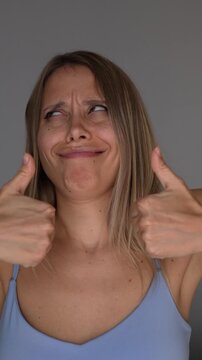 Young blonde woman gives a sarcastic thumbs-up with a skeptical facial expression, showing ironic praise, fake approval and passive-aggressive humor on a grey background