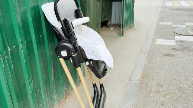 A lonely baby high chair stands abandoned against a green corrugated wall. It sits forgotten on a city sidewalk, evoking a sad and melancholic feeling of lost childhood.