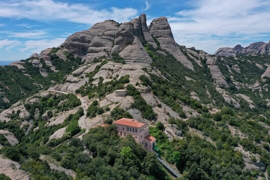 Aerial view of the Montserrat mountain range with its jagged rock formations, lush green vegetation, and the funicular station building in Monistrol de Montserrat, Catalunya, Spain.