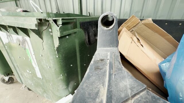 Broken green dumpsters sit piled with cardboard and waste. They rest neglected near a wall, creating a bleak and depressing atmosphere.