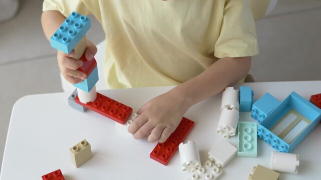 Child hands connect colorful construction pieces on white table creating simple structure. Educational activity demonstrates concentration, coordination and early engineering learning through play.