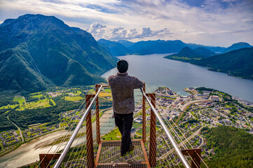 Naklejka premium Hiker on Nesaksla mountain, enjoying a scenic view of Andalsnes, Norway, and the surrounding Rauma River, Romsdalsfjord and steep Romsdal peaks under dramatic sky.