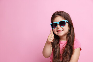 Fototapeta premium Cute little girl in sunglasses showing thumbs up on pink background, space for text