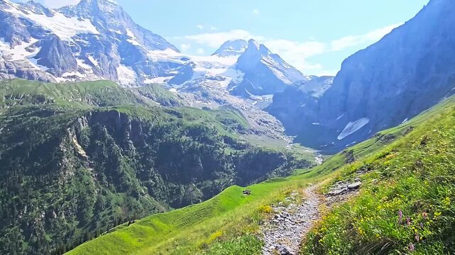 First-person perspective of hiking along a narrow rocky path on a steep green mountain slope, revealing a vast scenic valley and majestic snow-capped peaks under a sunny sky.