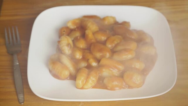 Plate of steaming hot potato gnocchi with tomato sauce on a wooden table, Freshly served Italian gnocchi pasta covered in savory red sauce with rising steam